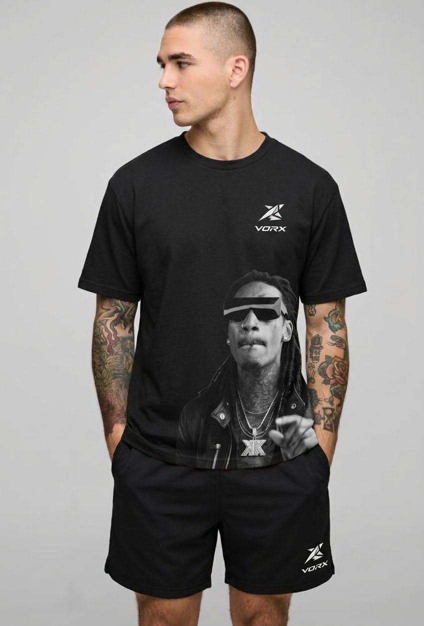 Man wearing black VORX t-shirt and shorts with tattoos on both arms, standing against gray background