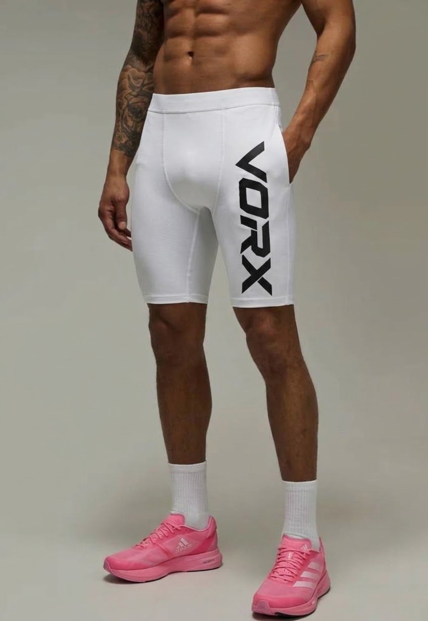 Shirtless man wearing white Vorx athletic shorts with black logo, white socks, and pink sneakers against a gray background
